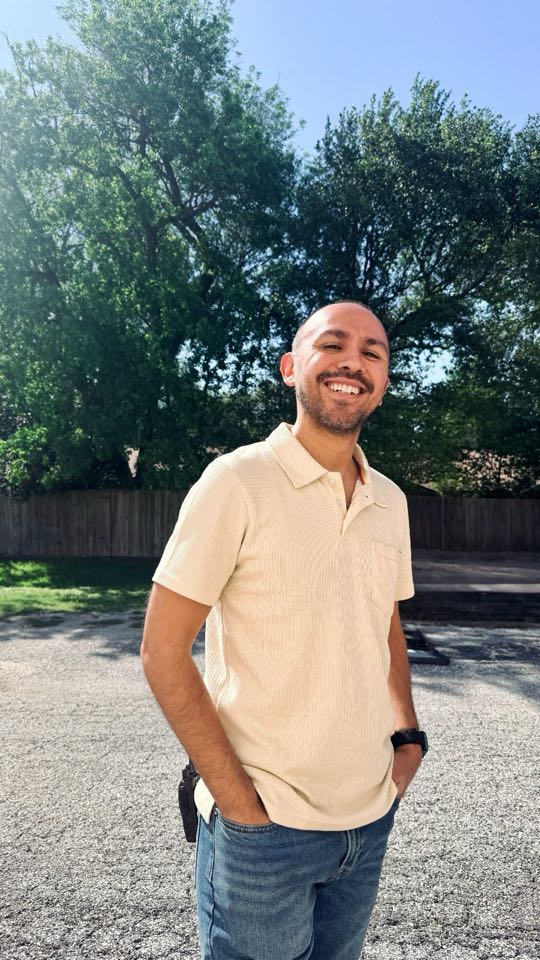 Nick grinning outdoors in a cream polo on a sunny day, trees behind him