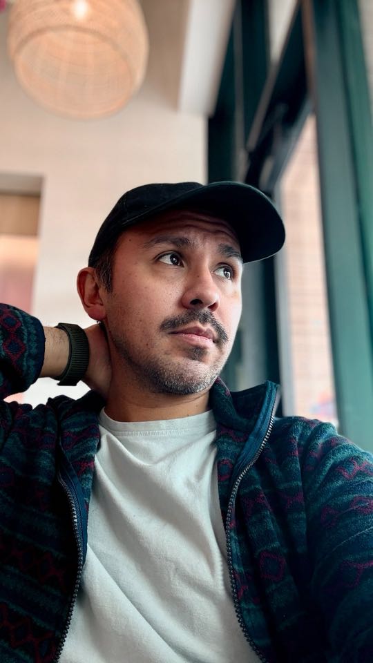 Nick in a black cap and patterned jacket, looking out a café window
